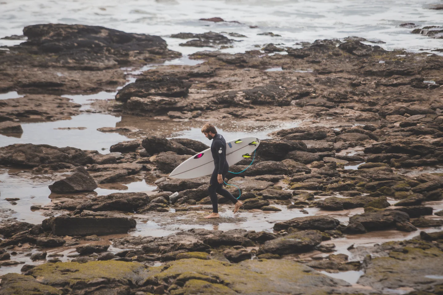 A surfer carrying a board across a rocky shoreline at Billabong Pro Miramar